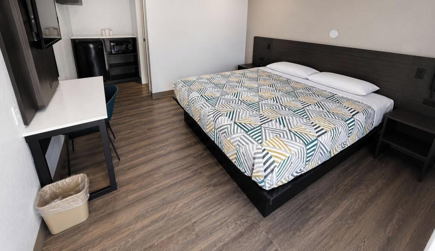 A hotel room with a bed, a desk, a chair, a trash bin, and a small kitchenette.