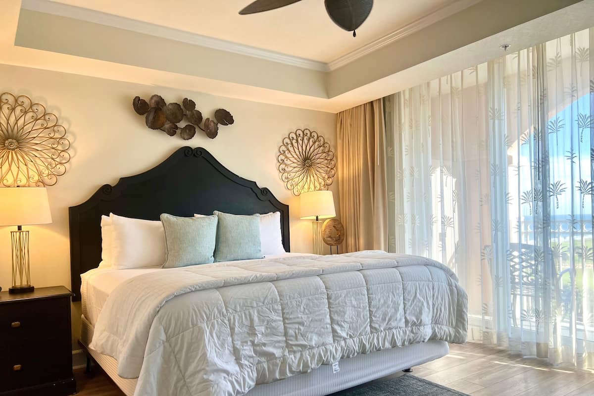 A spacious bedroom with a large bed, two bedside lamps, decorative wall art, and a view of the outdoors through sheer curtains.