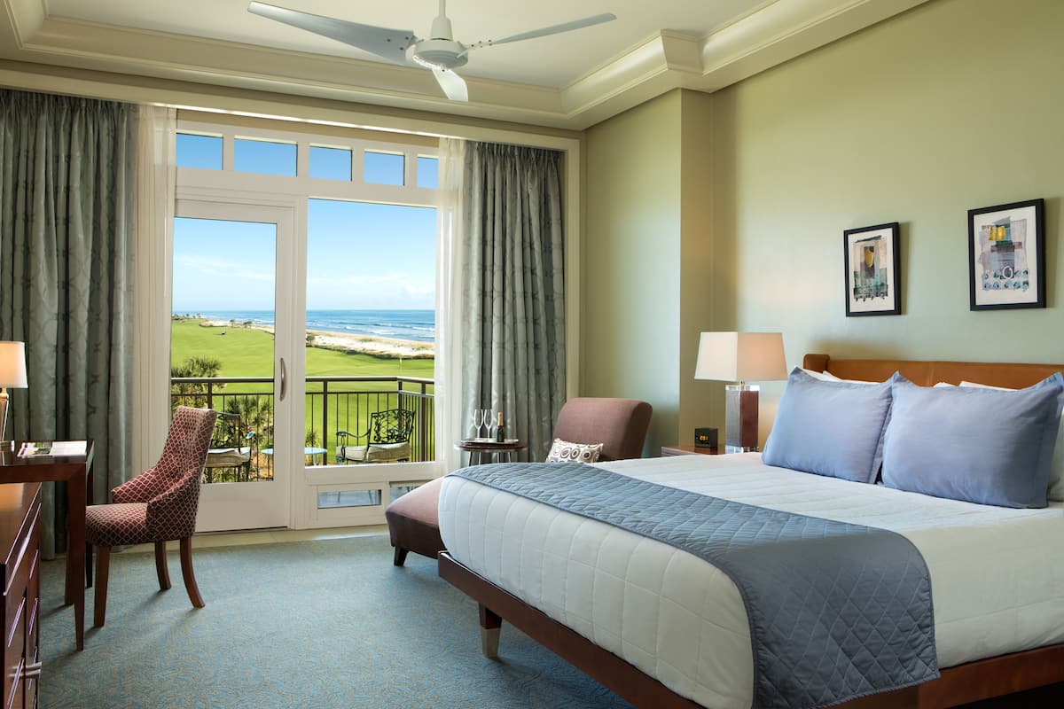 A hotel room with a large bed, a desk, a chair, and a balcony with a view of the ocean.