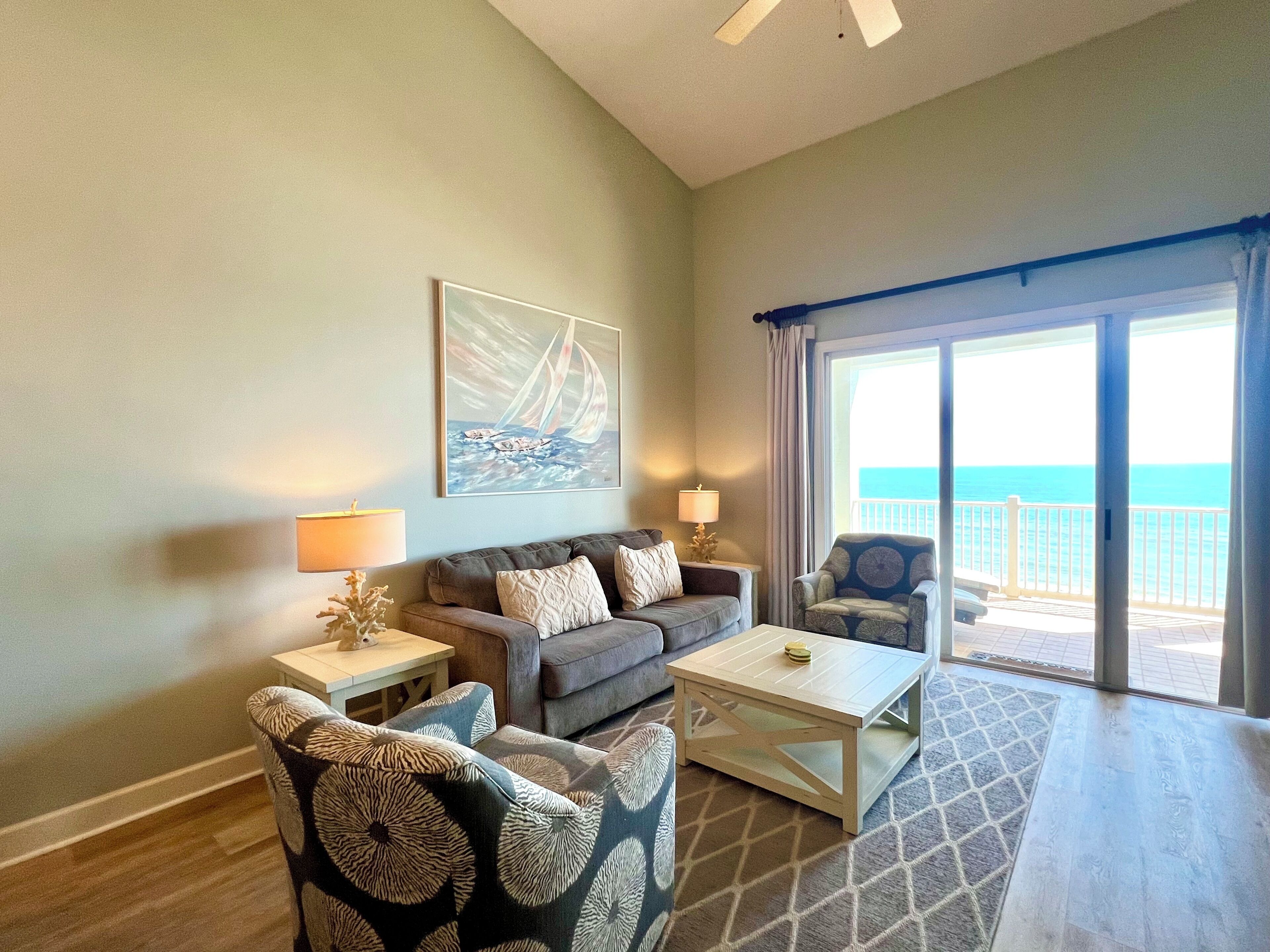 A living room with a sofa, armchairs, a coffee table, and a painting. There is a balcony with a view of the ocean.