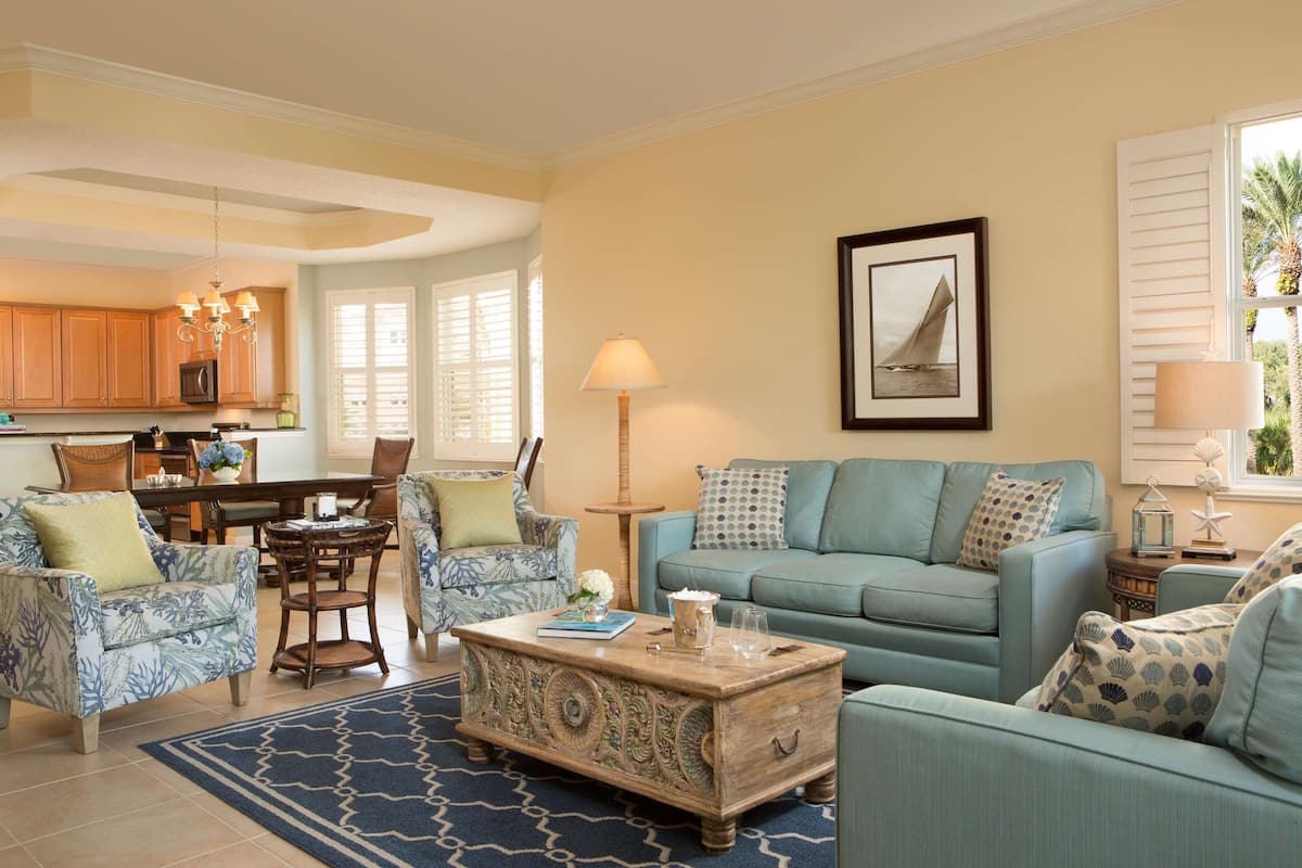 A living room with a light blue sofa, patterned armchairs, a coffee table, and a dining area with wooden cabinets and a chandelier.