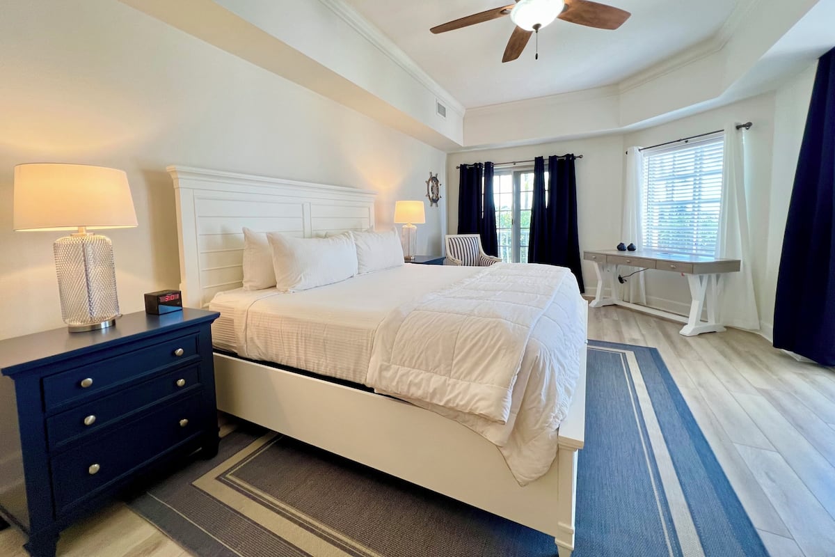 A bedroom with a bed, a nightstand, a desk, a chair, a ceiling fan, and a window with curtains.