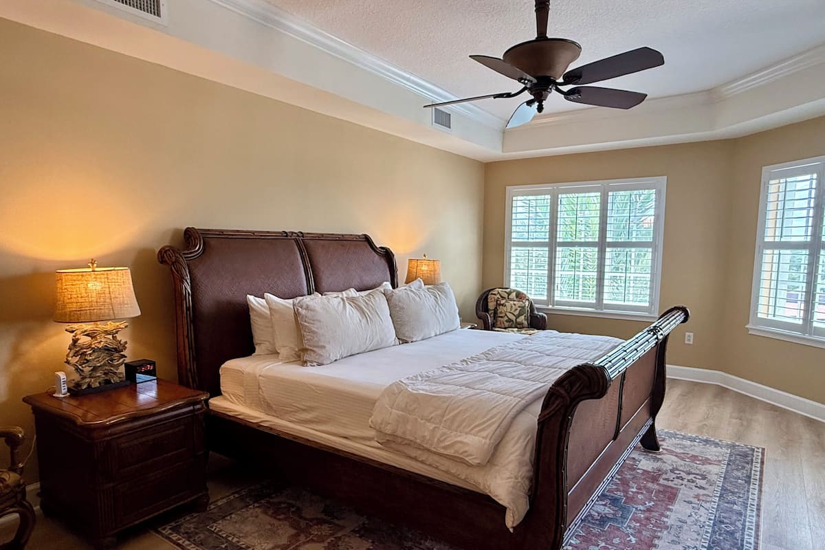 A bedroom with a large bed, a ceiling fan, a nightstand, a lamp, and a window with a view of greenery.