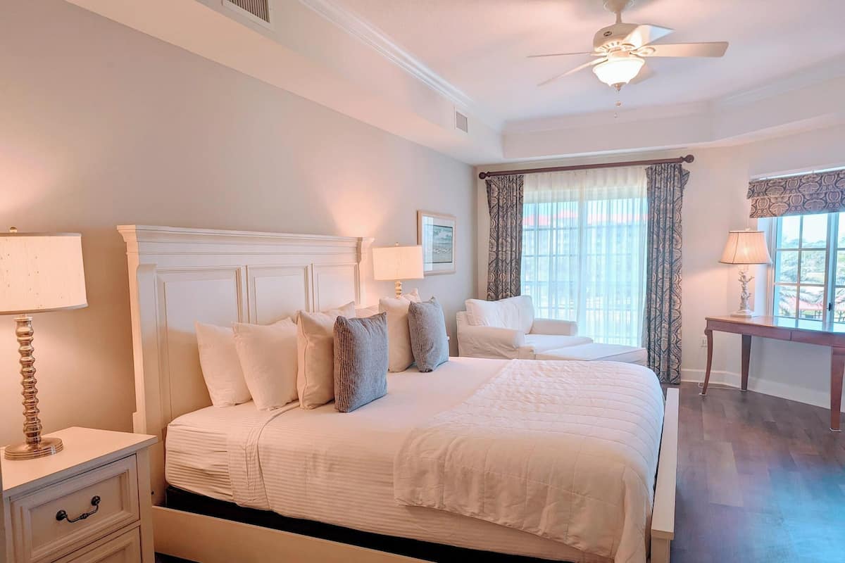 A bedroom with a large bed, a nightstand, a bedside table, a ceiling fan, and a window with curtains.