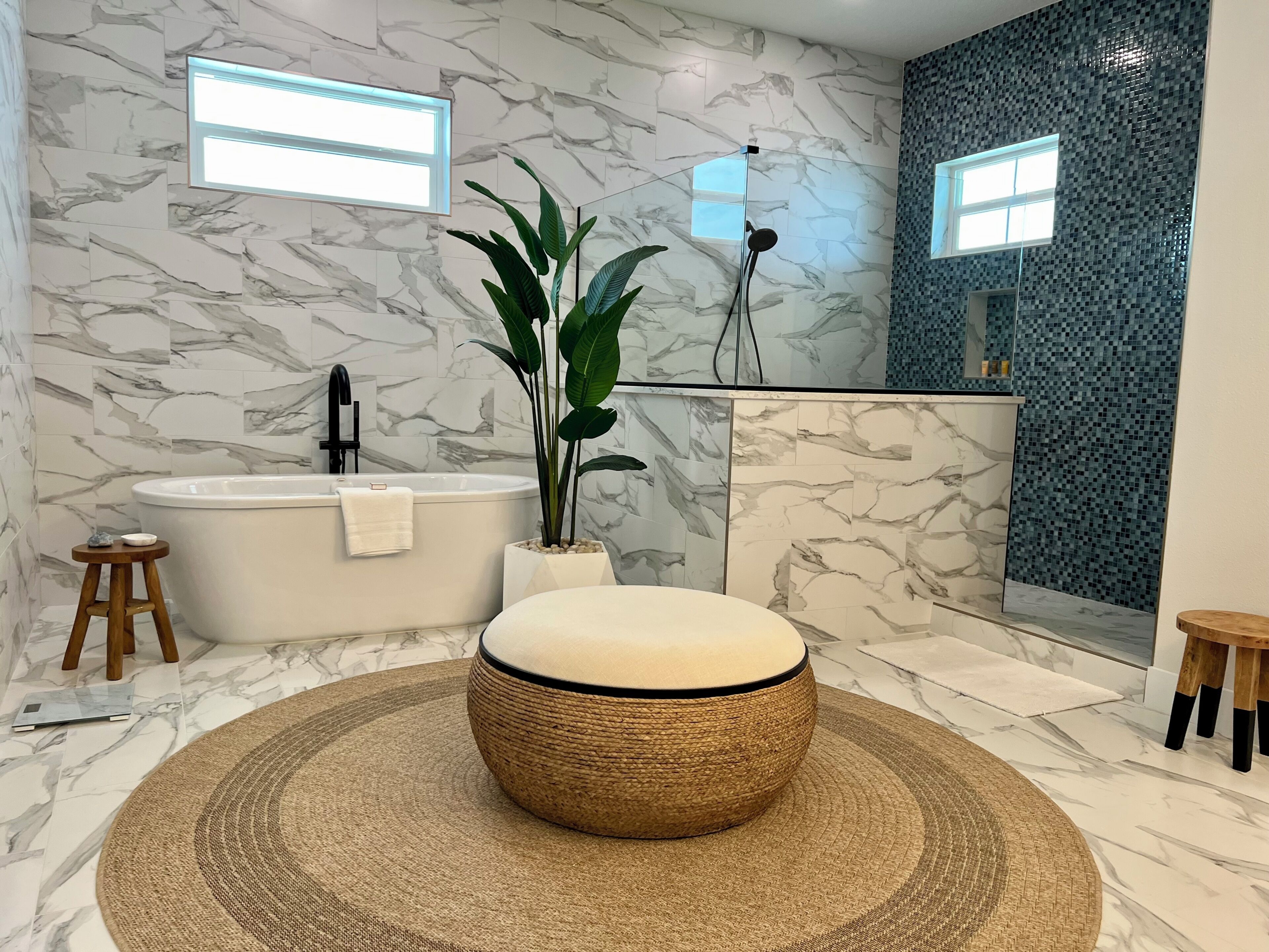 A modern bathroom with a freestanding bathtub, a large mirror, a plant, and a round ottoman.