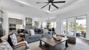 A spacious living area with a large sofa, coffee table, and dining table. There is a kitchen in the background with modern appliances.