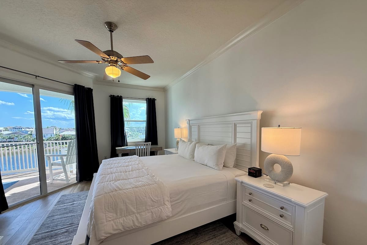 A hotel room with a large bed, a desk, a chair, a nightstand with a lamp, and a ceiling fan.