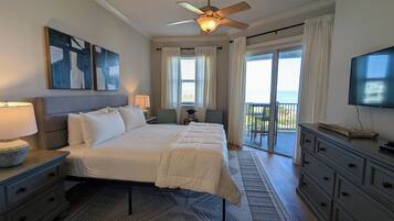 A bedroom with a bed, a ceiling fan, a TV, and a view of the ocean through a sliding glass door.