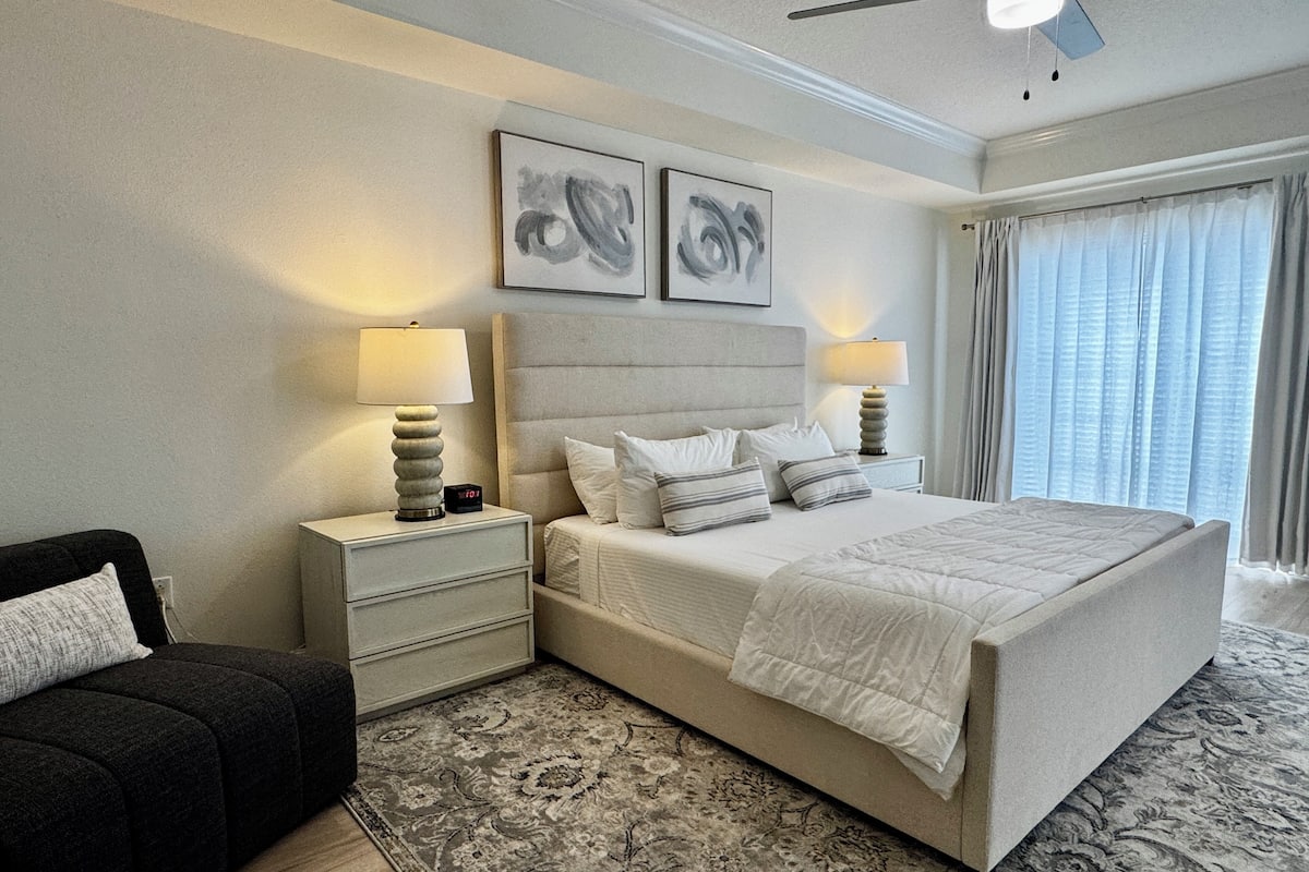 A bedroom with a large bed, two bedside tables with lamps, a black armchair, and a patterned rug.