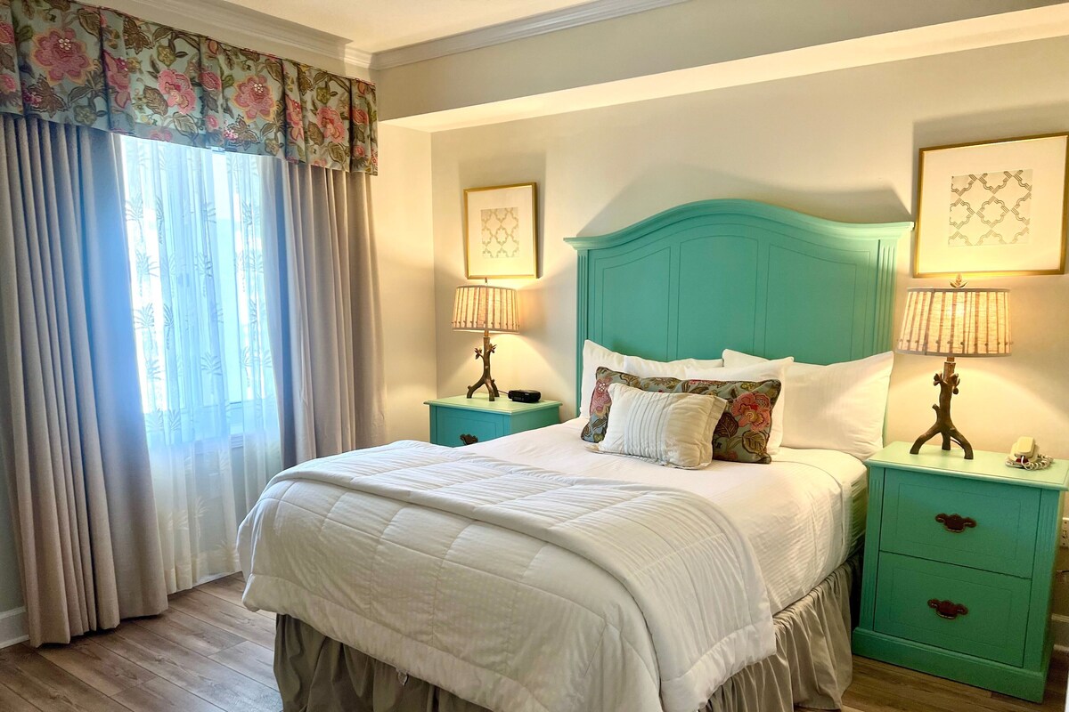 Una cama bien tendida con un edredón blanco y un cabecero verde aguamarina, flanqueada por mesitas de noche a juego con lámparas y una cortina con estampado floral.