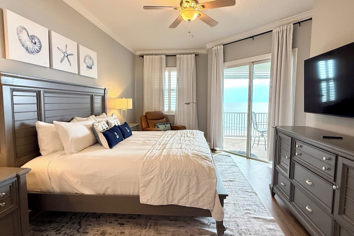 A hotel room with a bed, a TV, a ceiling fan, a chair, and a view of the ocean.