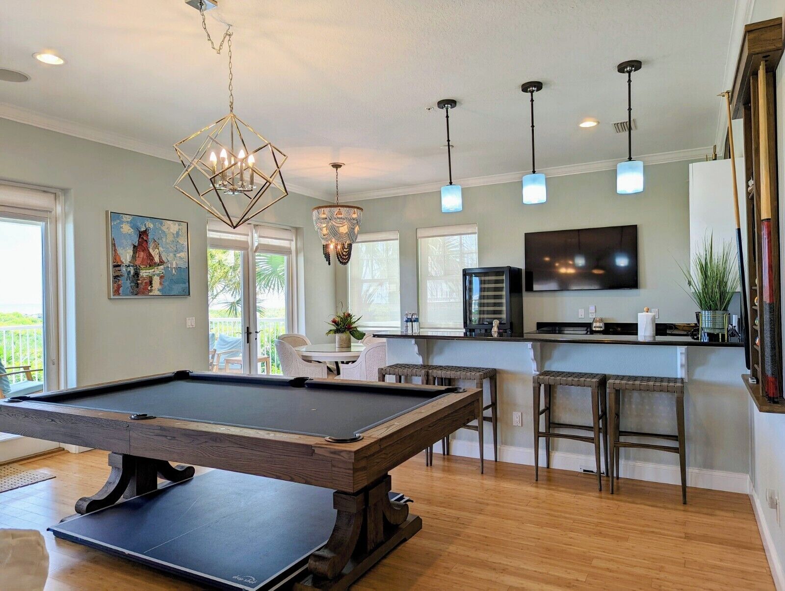 A pool table in a room with a bar area, pendant lights, a painting, and a television.