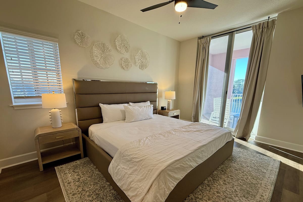 A bedroom with a large bed, two bedside tables, a ceiling fan, a window with blinds, and a view of the outside.