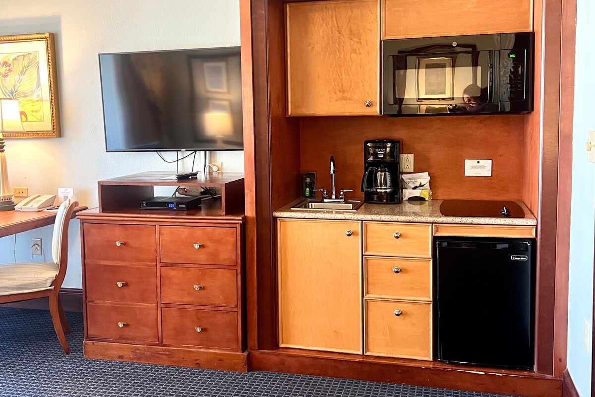 Habitación de hotel compacta con un mueble de madera que contiene un microondas, una cafetera y un refrigerador pequeño. Un televisor de pantalla plana está montado en la pared sobre el mueble.