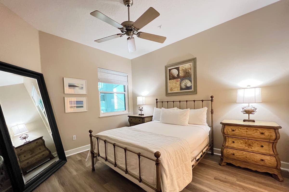 A bedroom with a bed, a dresser, a ceiling fan, a window with blinds, and framed artwork on the wall.