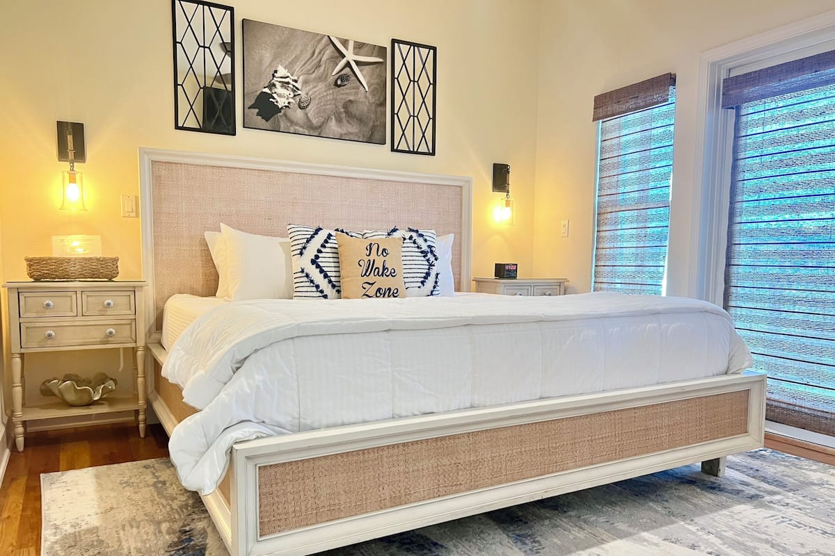 A bedroom with a large bed, a bedside table, a nightstand, wall art, and a window with blinds.