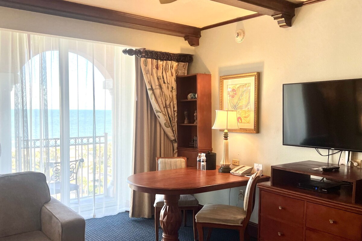 Habitación de hotel con un ventanal grande, una mesa de madera, un televisor, un sofá y un escritorio con cajones.
