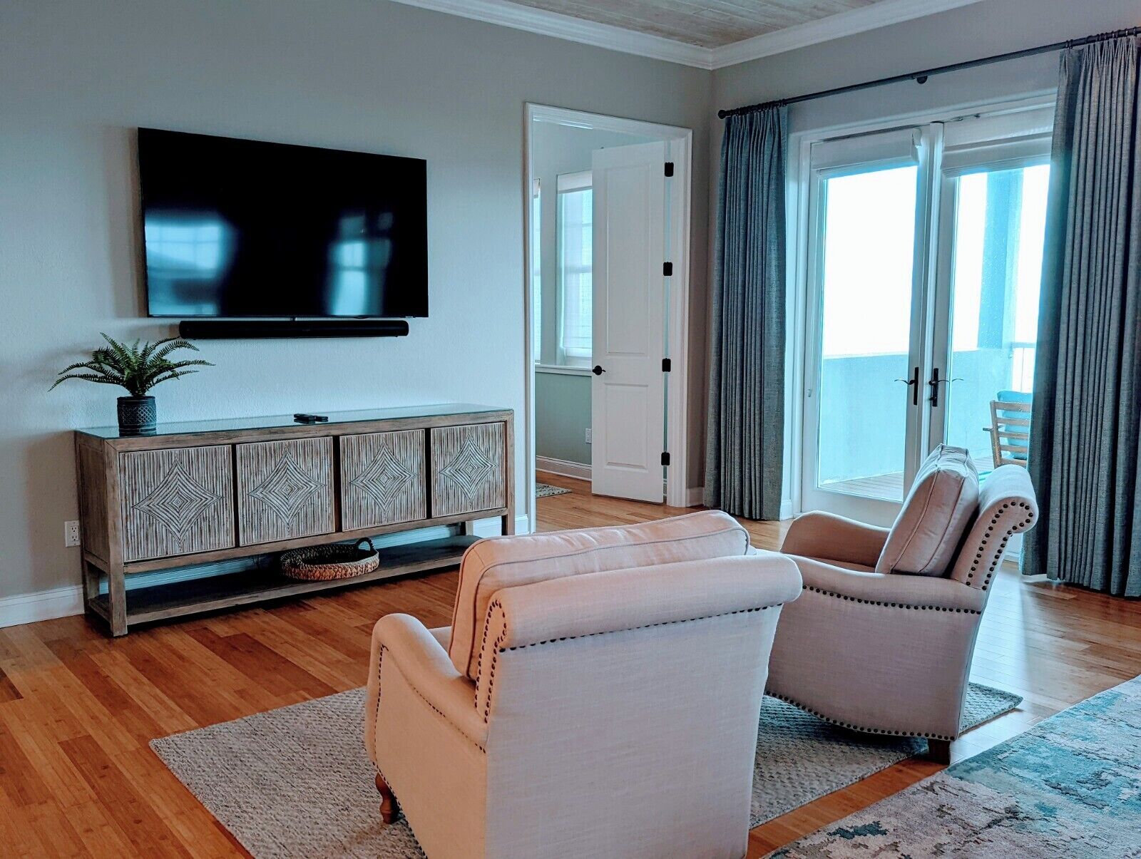 Una sala de estar con una televisión de pantalla plana, un aparador de madera con diseños geométricos, dos sillones color beige, una alfombra estampada y un ventanal corredizo que da a un balcón.