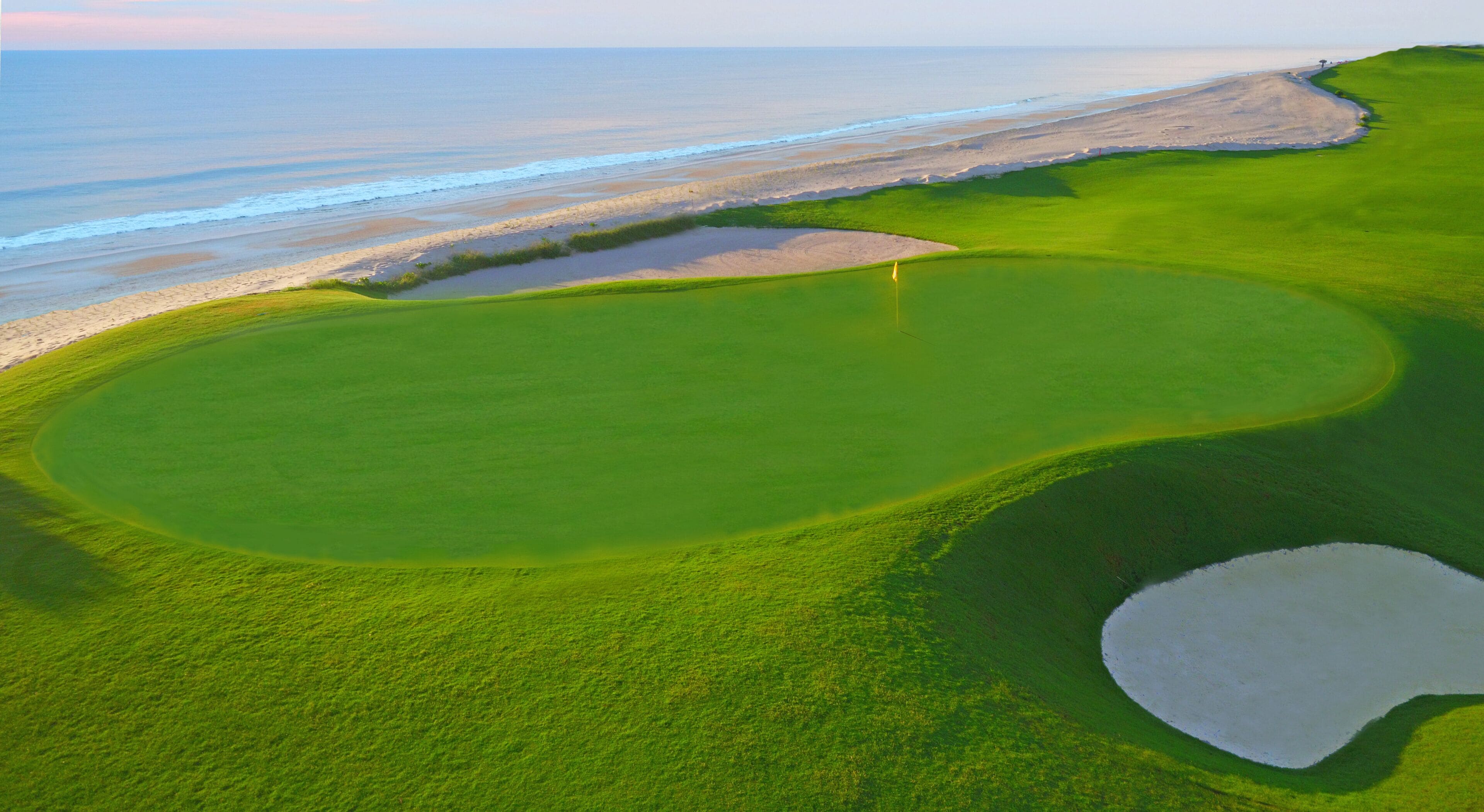 A golf course with a large green, a water hazard, and a beach in the background.