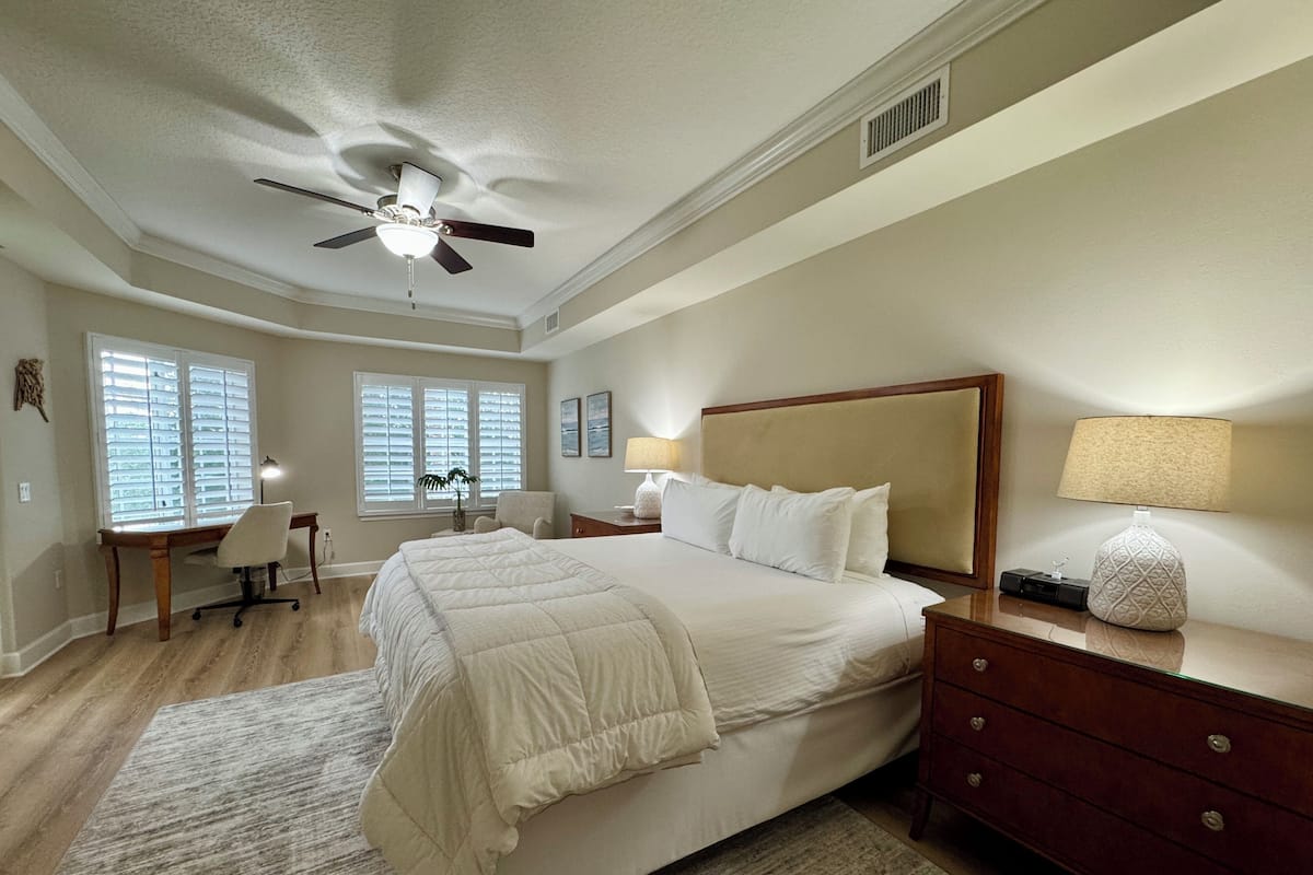 A bedroom with a bed, a desk, a chair, a nightstand, a lamp, and a ceiling fan.