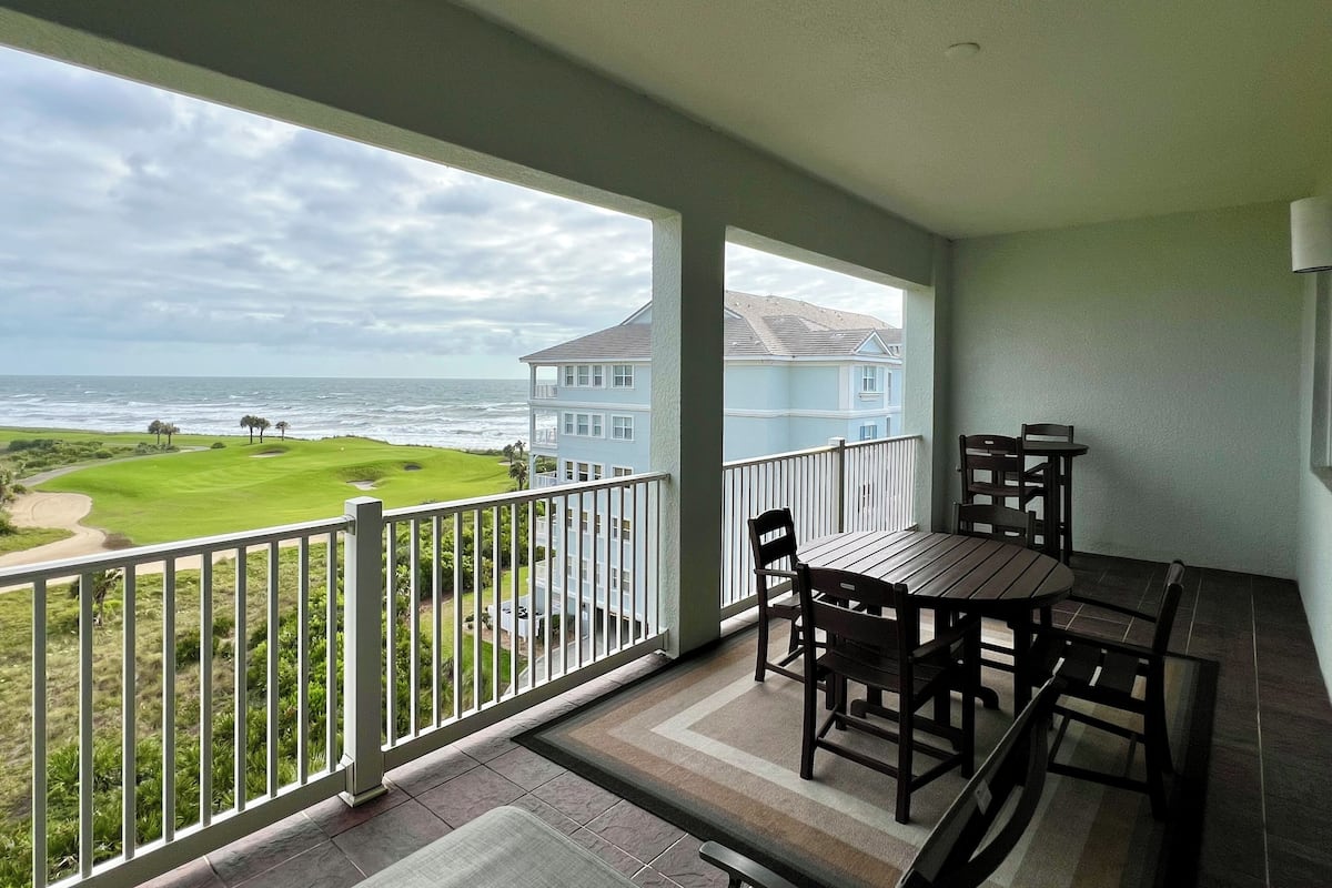 A balcony with a view of the ocean and a golf course, a dining table with chairs, and a light fixture on the wall.