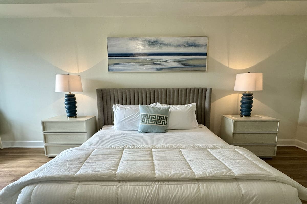 A bedroom with a large bed, two bedside tables, a headboard, and a painting on the wall.