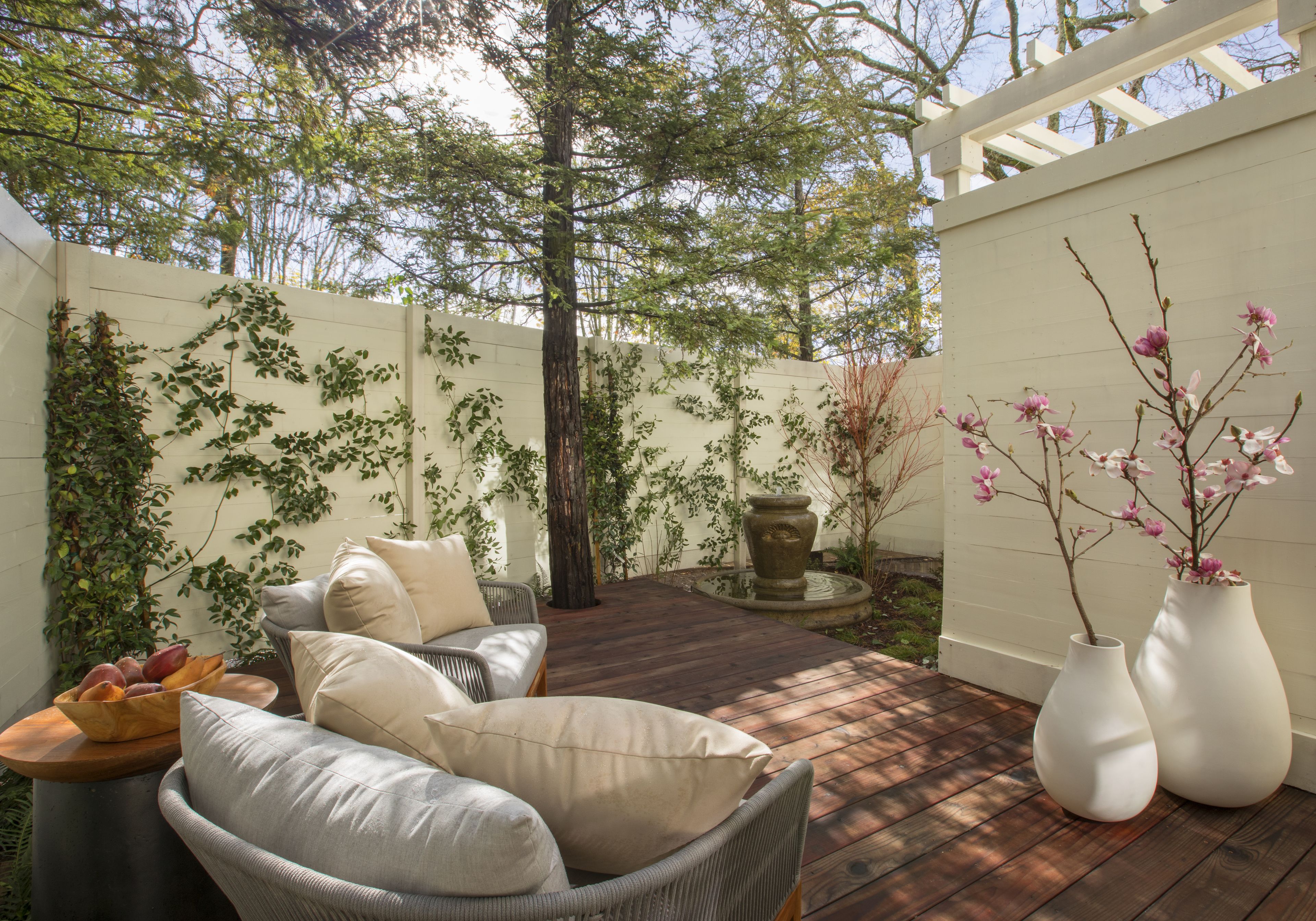 Un patio con mobiliario de mimbre, una mesa de madera con fruta y dos jarrones blancos con flores.