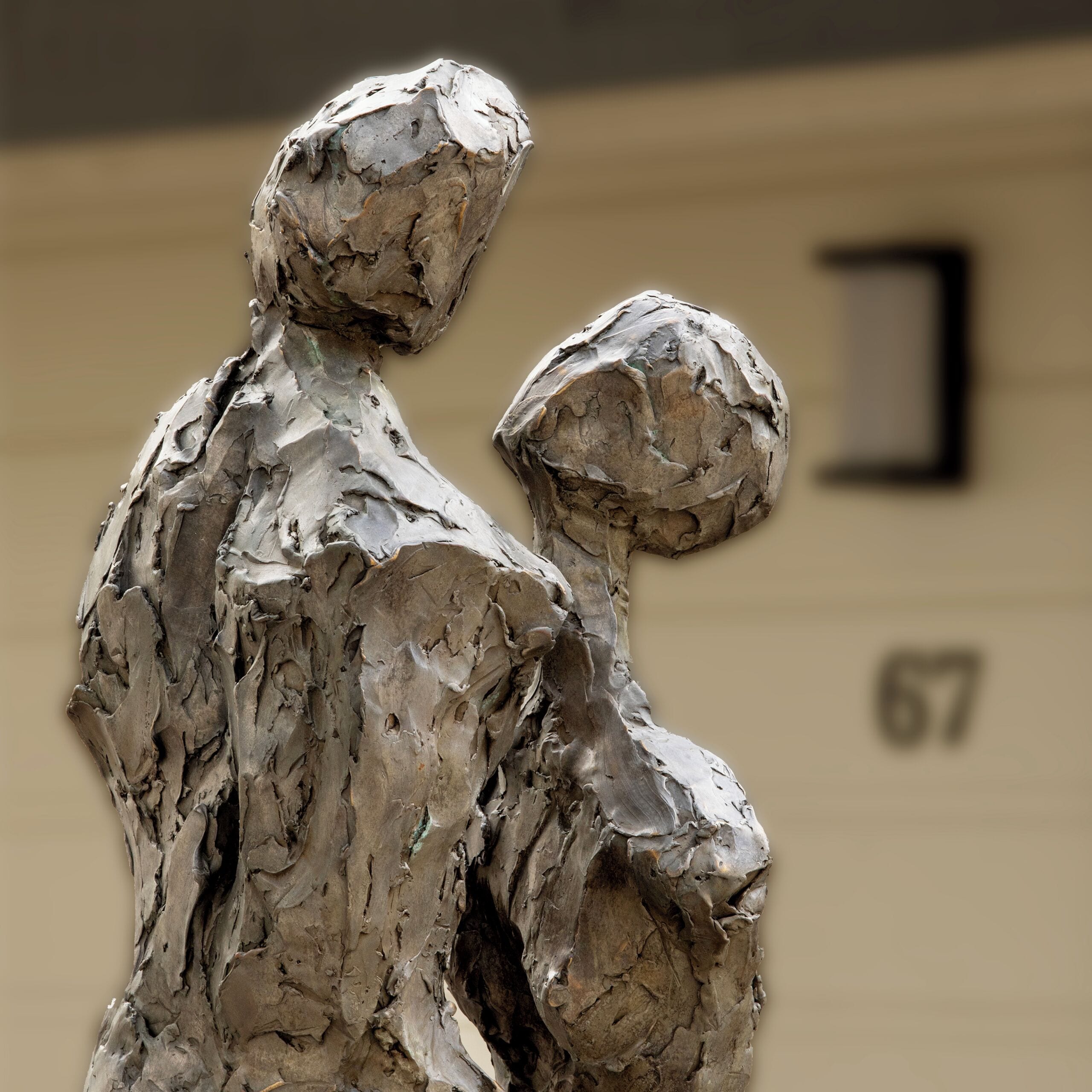 Una escultura de dos figuras, una más grande y otra más pequeña, con un fondo desenfocado que muestra una parte de un edificio con el número "67".