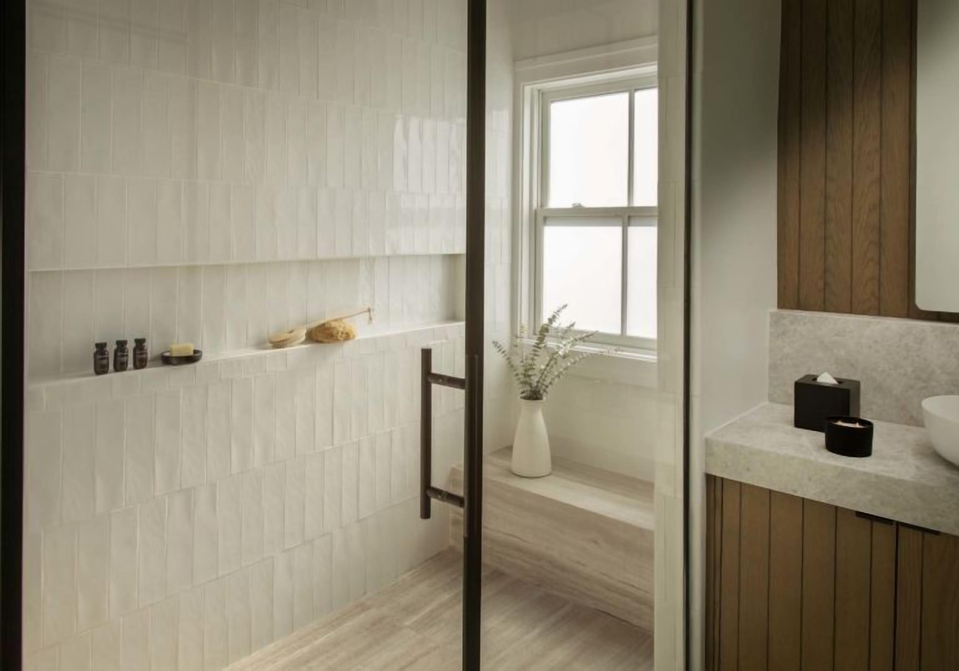 A bathroom with a white tiled wall, a wooden floor, a window, and a mirror above the sink.
