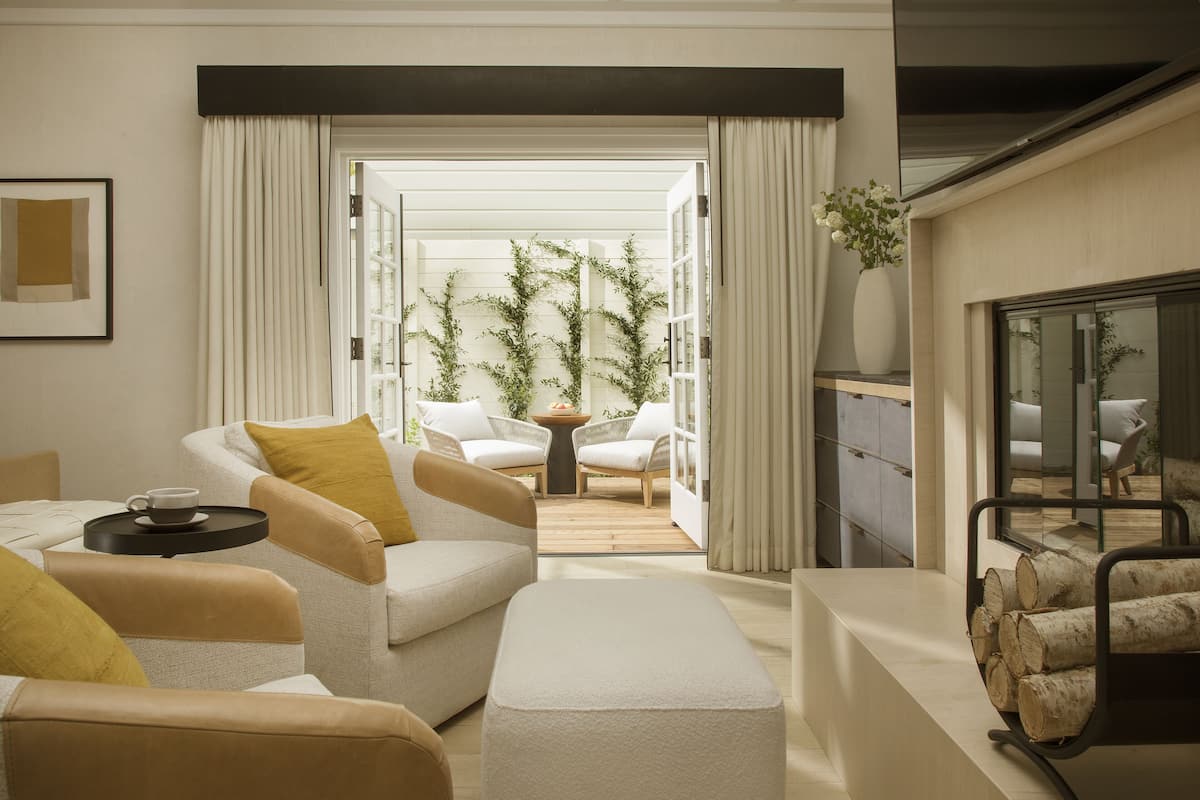 A modern living room with a sofa, ottoman, and a coffee table. There is a fireplace with logs, a side table with a vase, and a view into a balcony with plants.