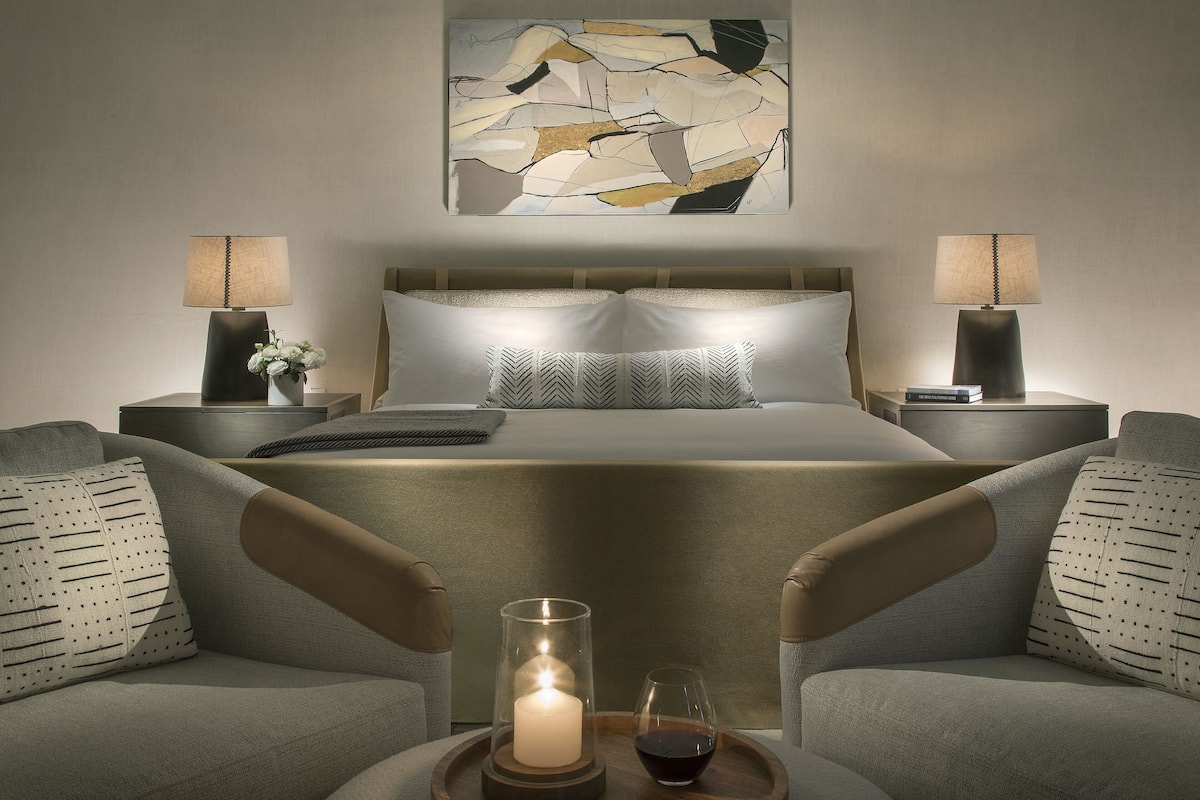 A modern hotel room with a large bed, two side tables with lamps, a decorative wall piece, and a small table with a candle and wine glass.