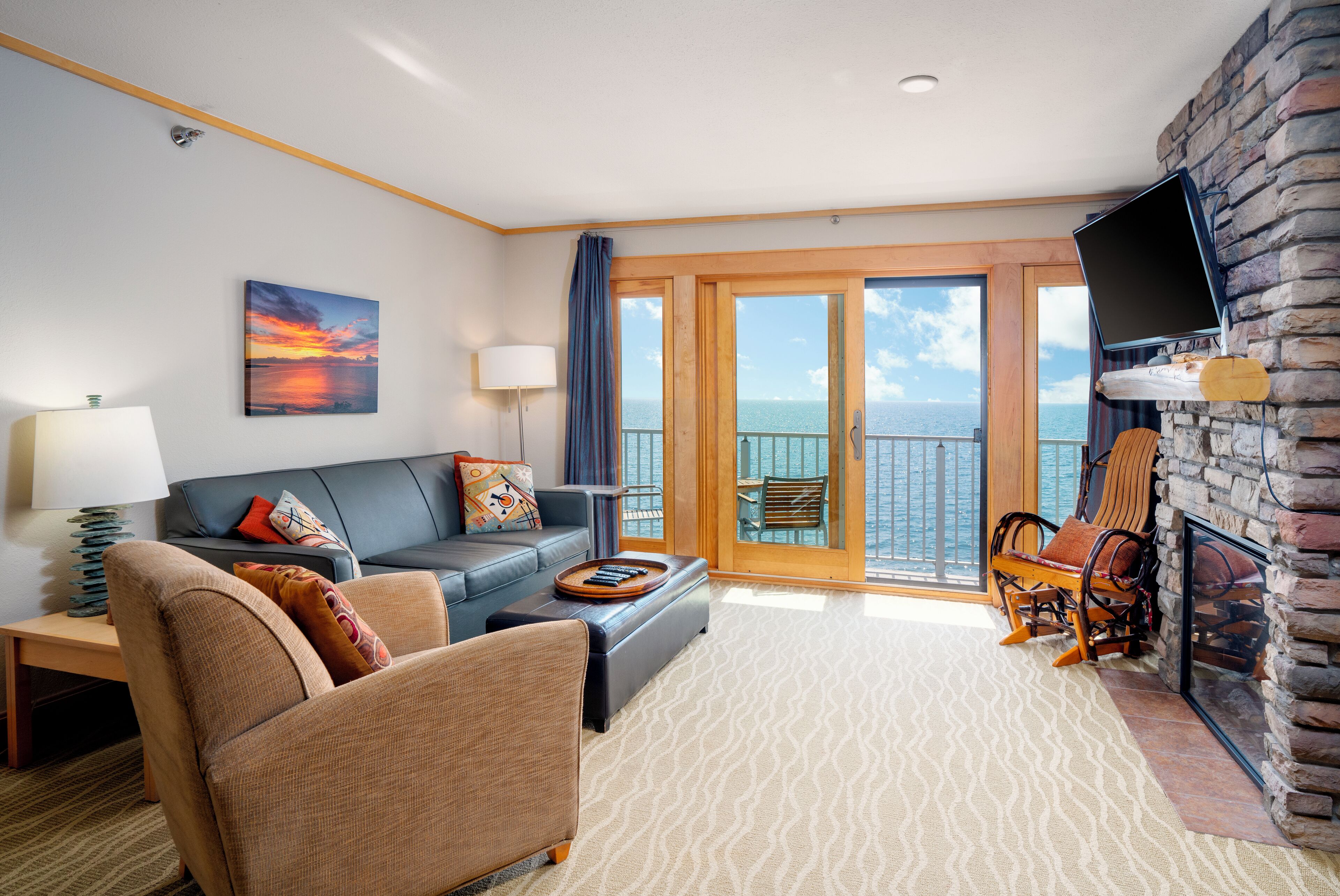 A living room with a sofa, armchair, and a coffee table. There is a television mounted on a stone wall and a balcony with a view of the sea.