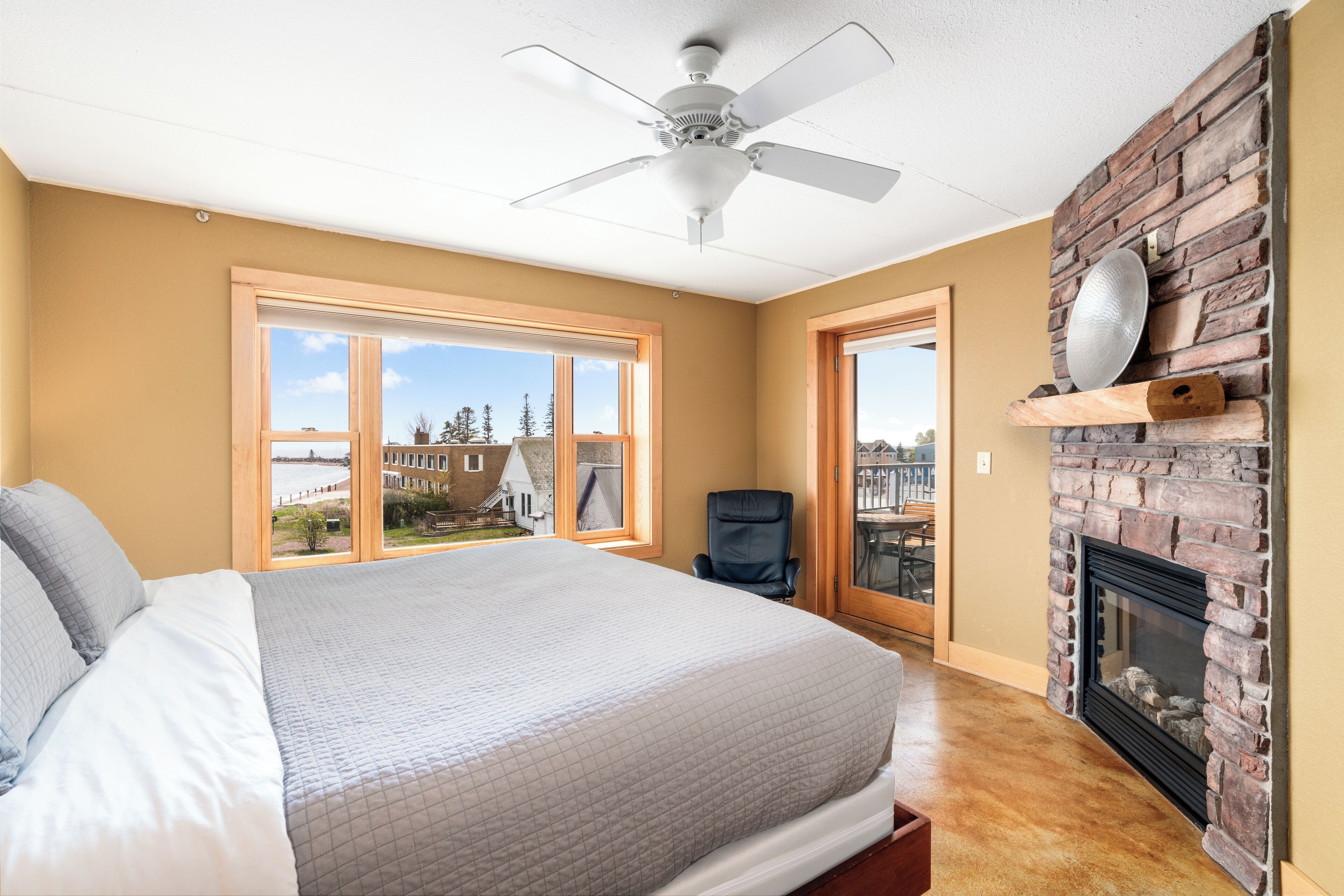 A bedroom with a large bed, a ceiling fan, a fireplace, and a view of the outside.