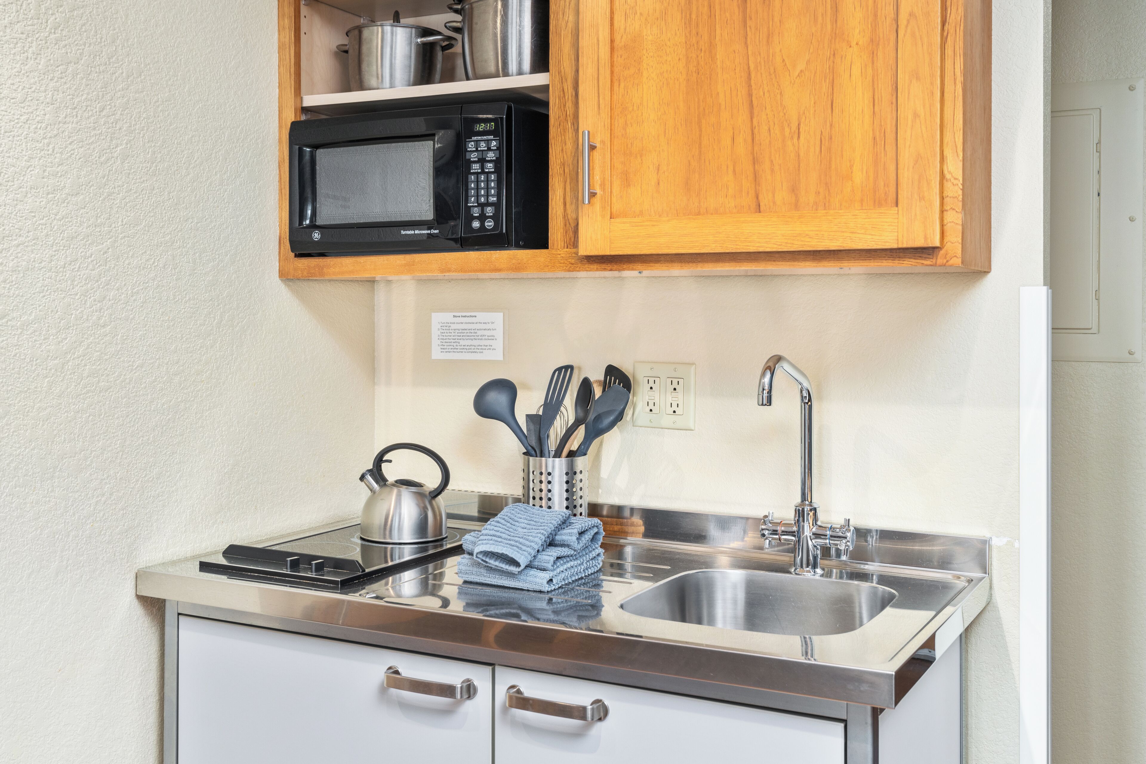 A compact kitchen with a microwave, kettle, sink, and stainless steel cabinets.