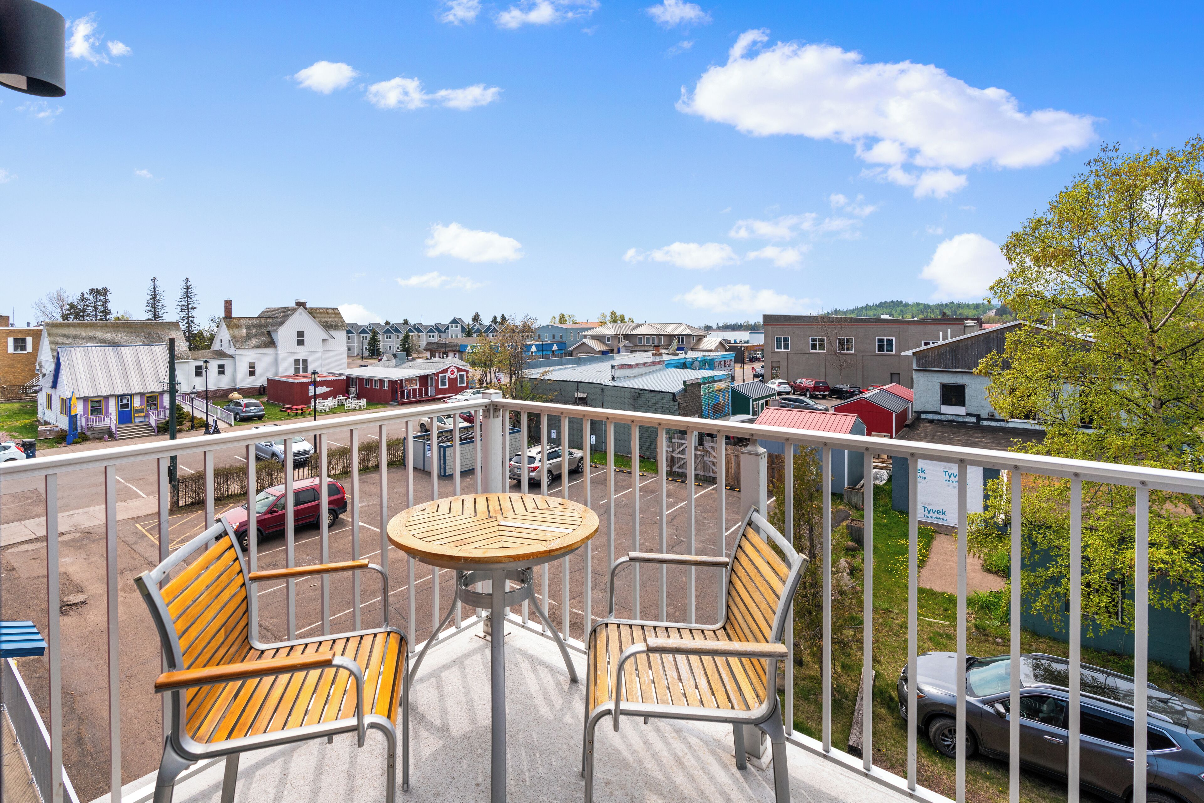 Ein Balkon mit zwei Holzstühlen und einem kleinen Tisch, mit Blick auf einen Parkplatz und Gebäude.