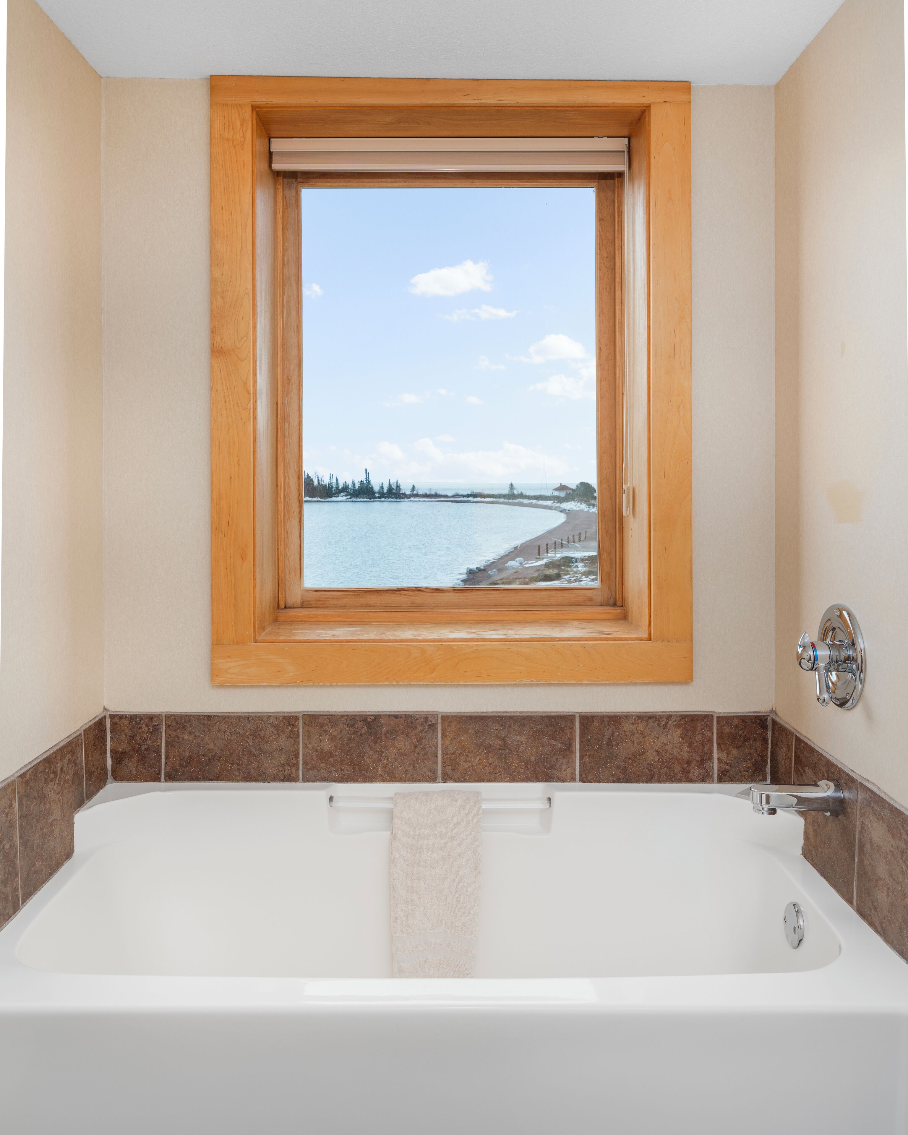 Un baño con una bañera grande, una ventana que ofrece vista a una playa y árboles, y paredes con azulejos marrones.