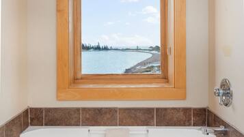 Un baño con una bañera grande, una ventana que ofrece vista a una playa y árboles, y paredes con azulejos marrones.