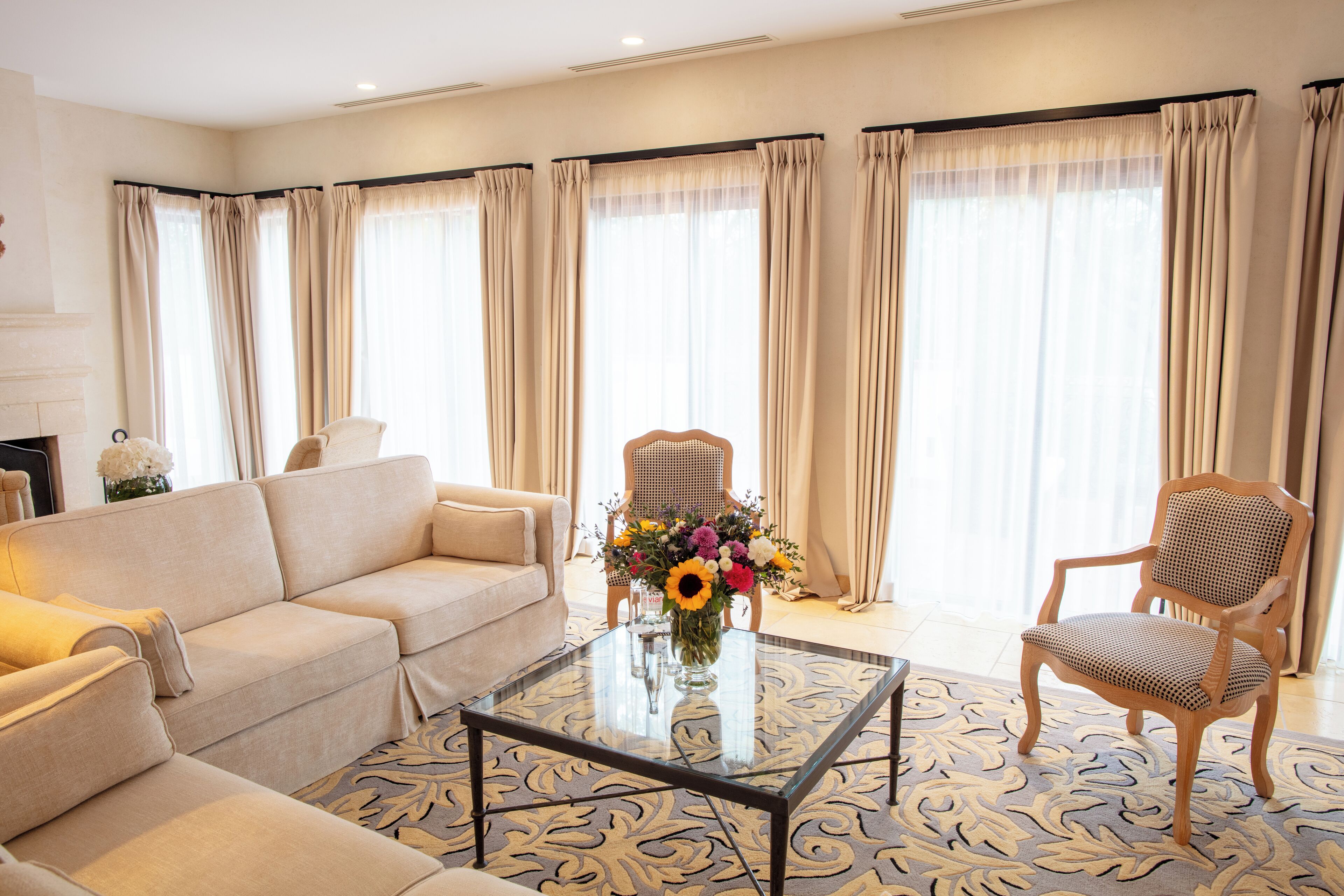 A spacious living room with a beige sofa, a wooden chair, and a glass coffee table with a floral centerpiece.
