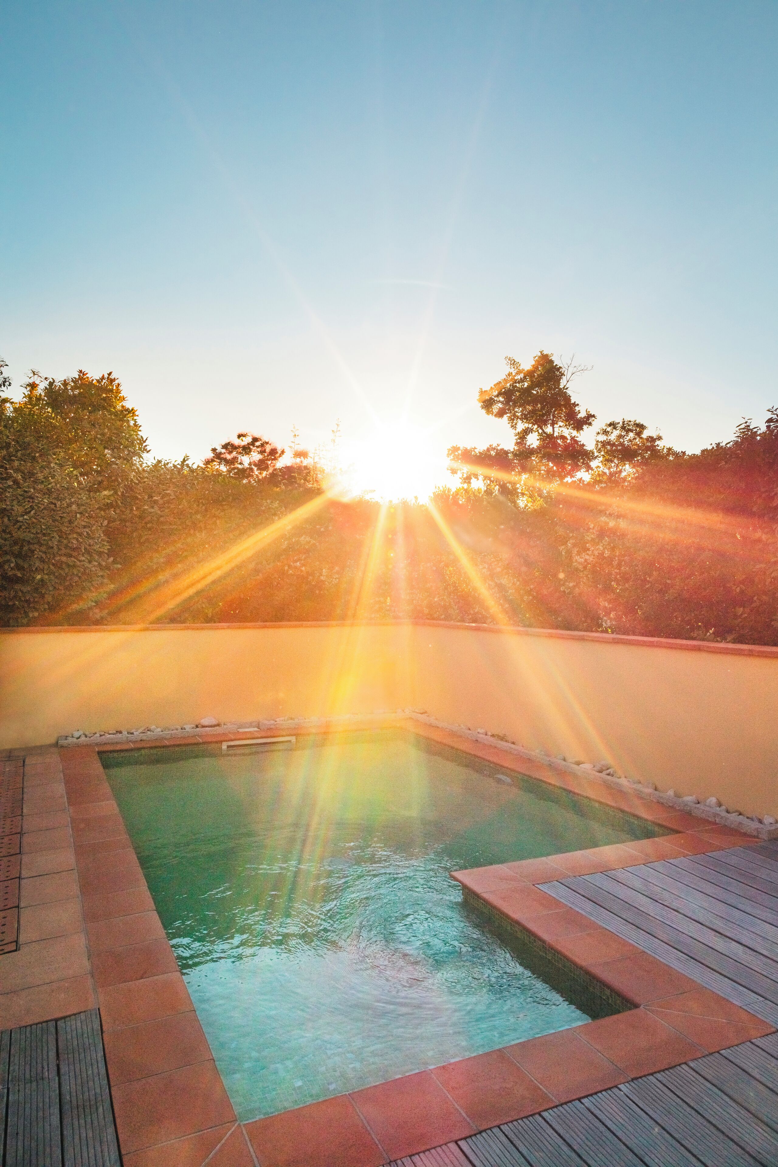 Una piscina con bordo in legno, circondata da un muretto basso e alberi sullo sfondo.