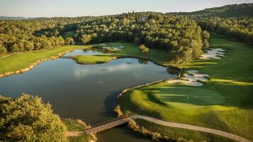 Stroll across the serene golf course, surrounded by lush greenery and rolling hills, with a picturesque pond and a bridge offering access across the water.