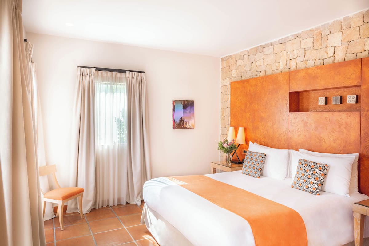 Habitación de hotel con una cama grande, una silla de madera, un muro de piedra y una ventana con cortinas.
