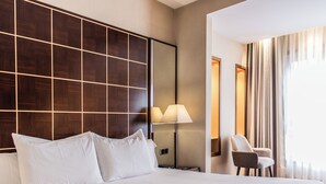 Habitación de hotel con una cama grande, una mesita de noche, una lámpara, una silla y un ventanal con cortinas.