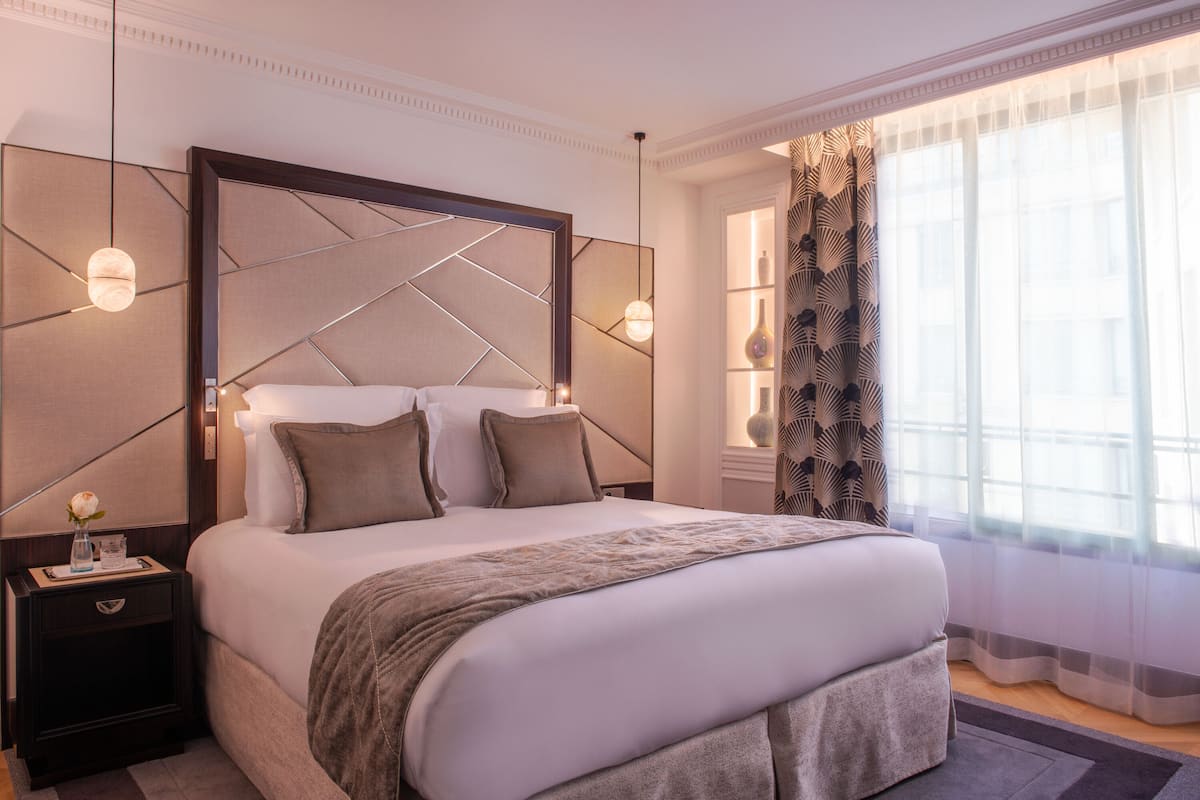 Una habitación de hotel moderna con una cama grande, una mesita de noche y un ventanal con cortinas.