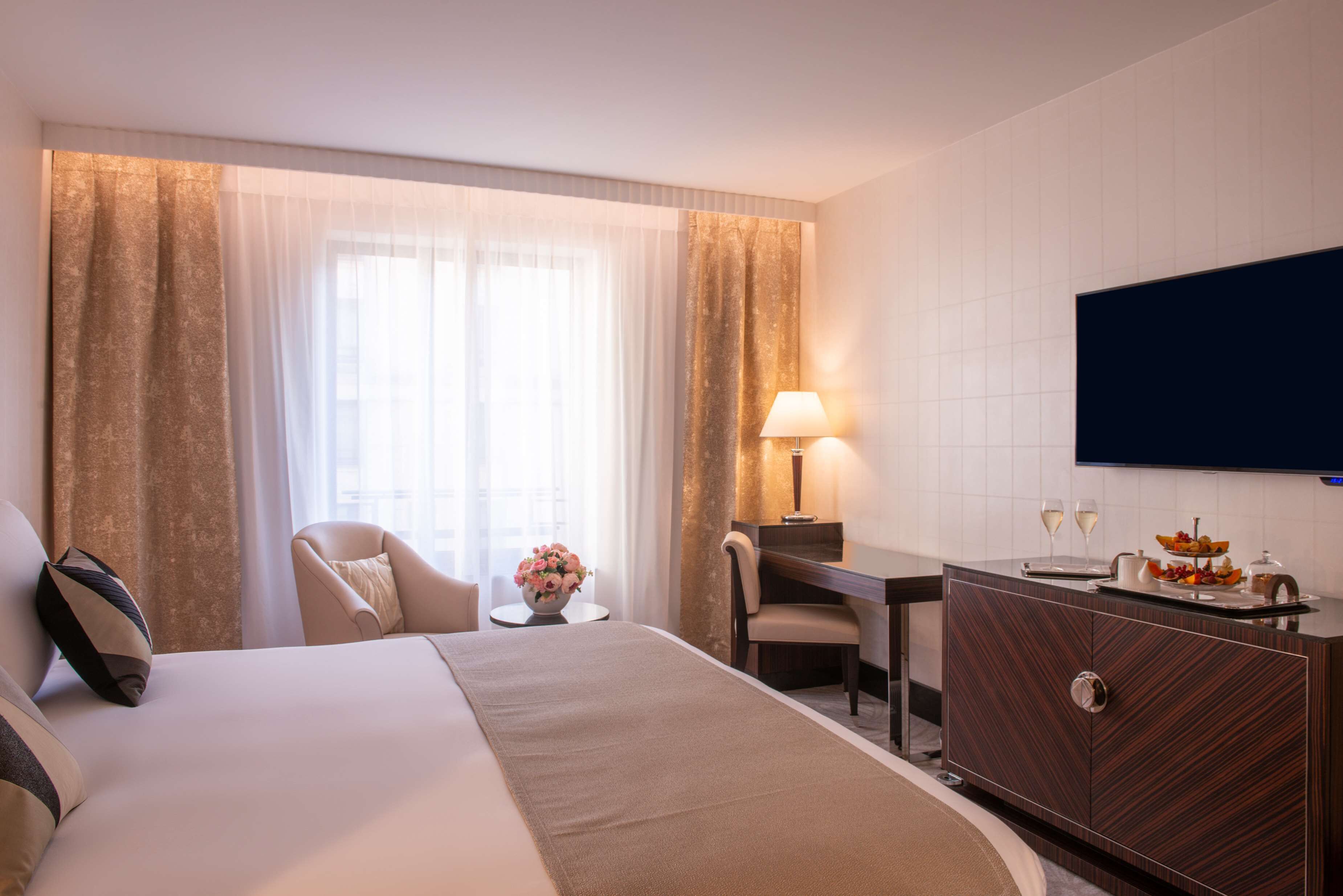 A hotel room with a bed, a desk, a chair, a TV, and a minibar.