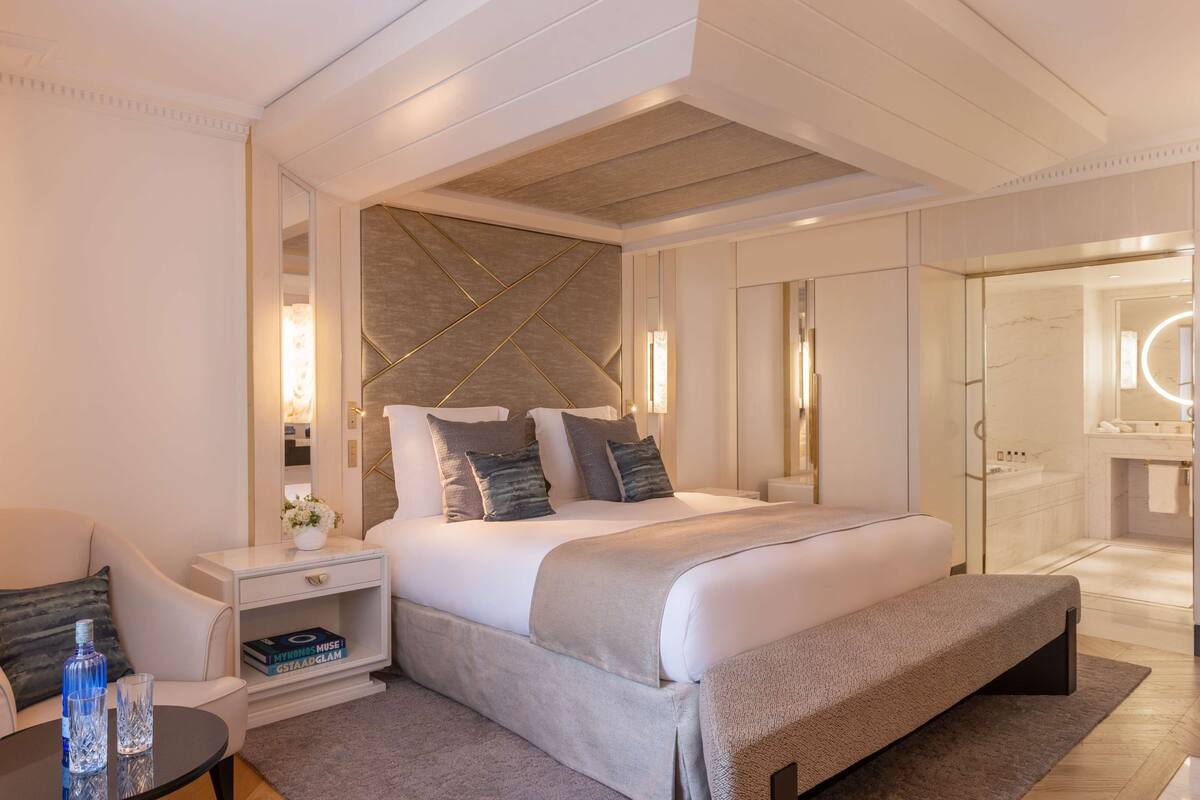Una habitación de hotel moderna con una cama grande, mesitas de noche, un banco y un baño con espejo y lavamanos.