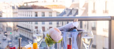 A table with a fruit platter, a pitcher, a teapot, a cup, and a dessert under a glass dome, set on a balcony with a city view.