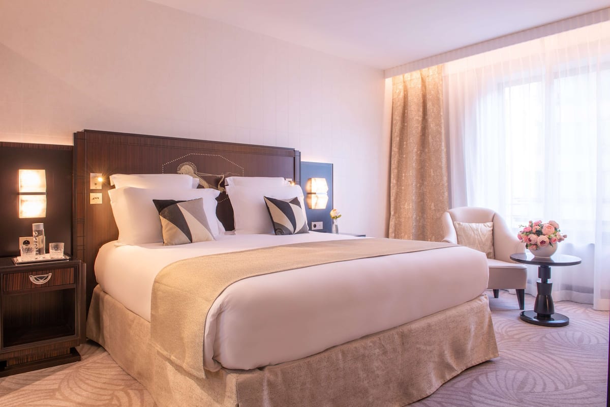 Habitación de hotel con una cama grande, una mesita de noche con lámpara, una silla y una pequeña mesa con un jarrón de flores.