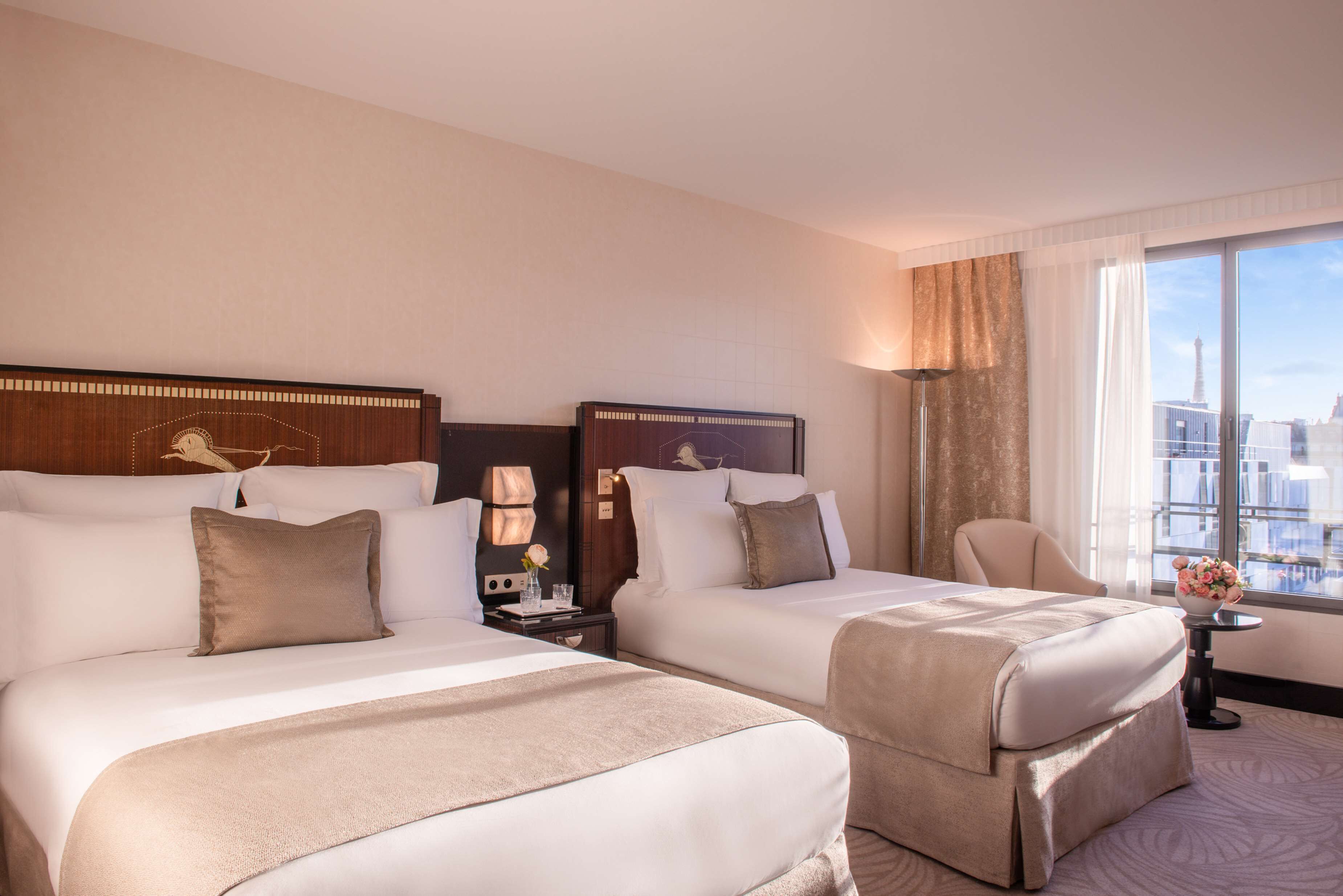 A hotel room with two beds, a nightstand with a lamp, a chair, and a view of the city.
