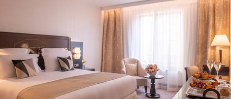 A hotel room with a large bed, a desk with a lamp, a chair, and a table with fruit and glasses.