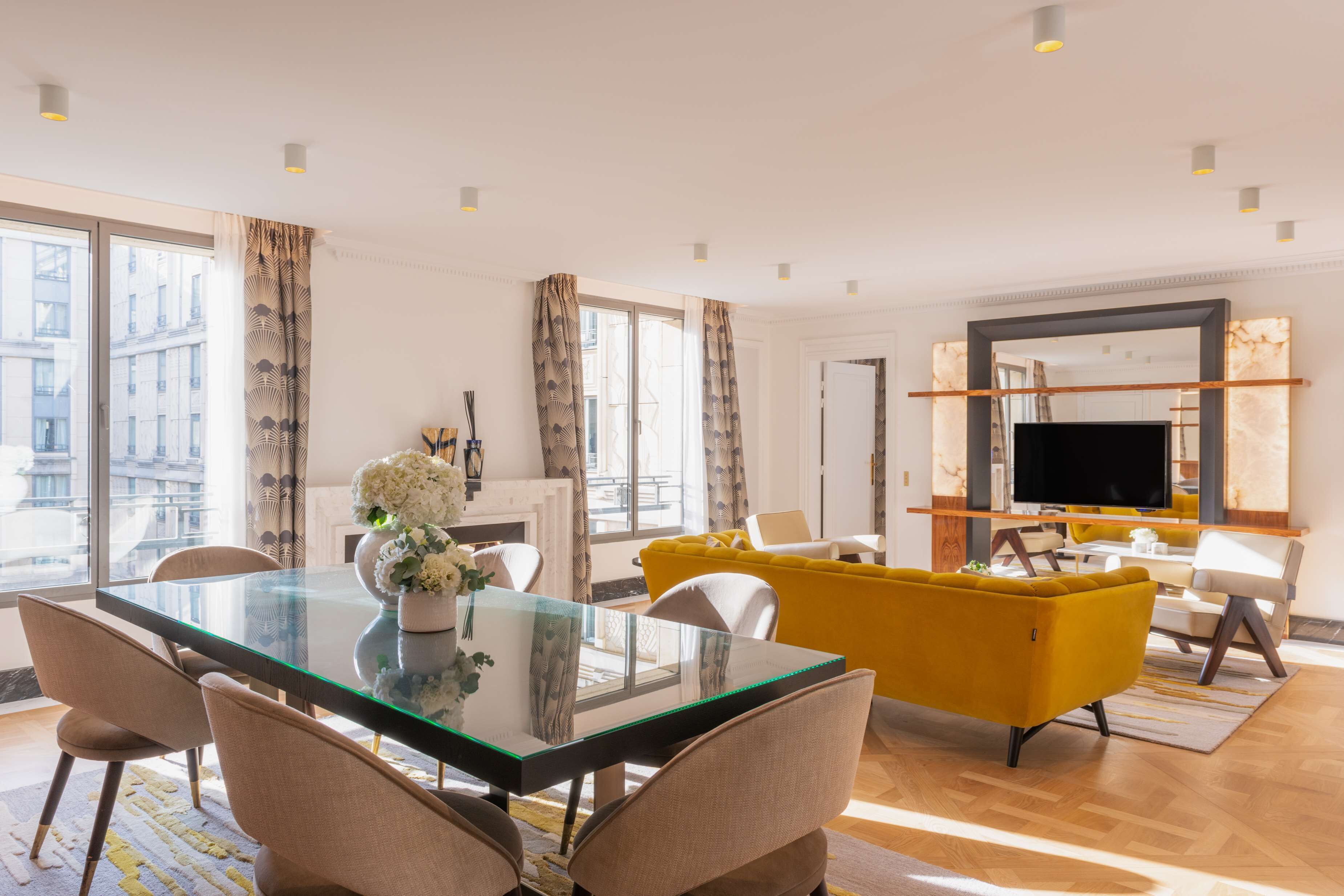 A modern living room with a glass dining table, yellow sofa, and a large TV.